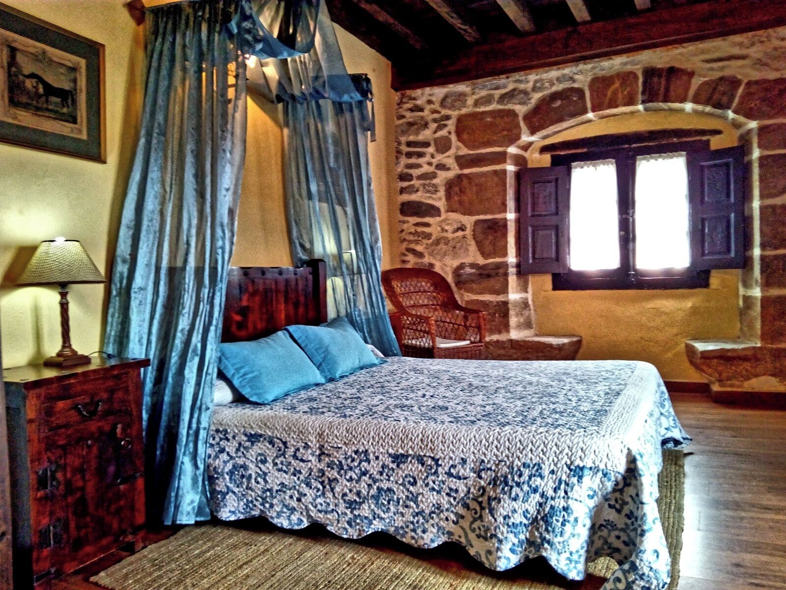 Un dormitorio con pared de piedra, una cama con colcha estampada, una mesita de noche de madera, una lámpara y una silla.
