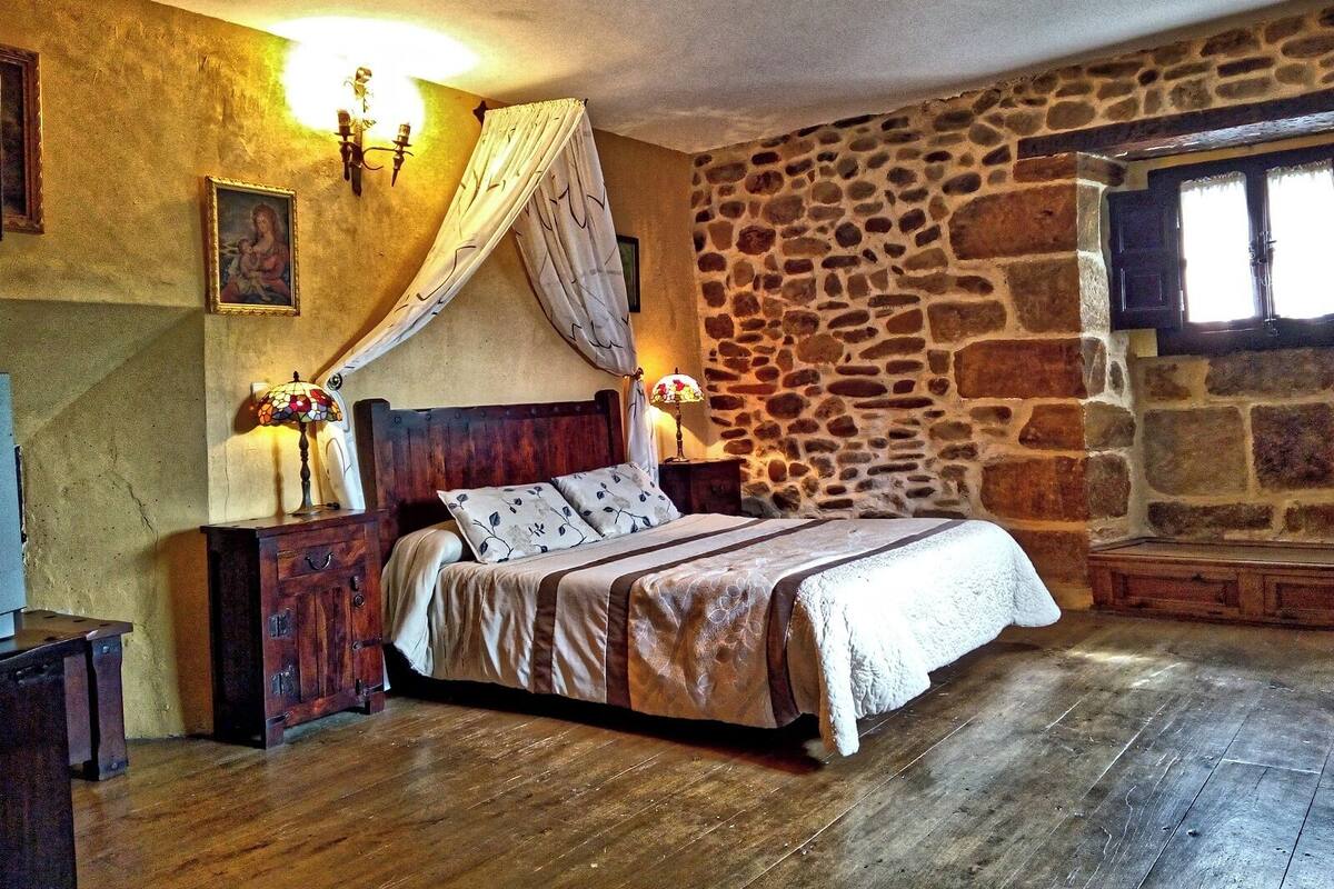 A bedroom with a stone wall, a wooden bed, a nightstand, a lamp, and a window with curtains.