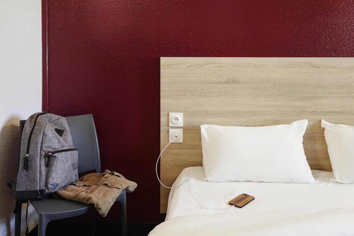 Una habitación de hotel con una cama, una silla, una mochila y un teléfono sobre la cama.