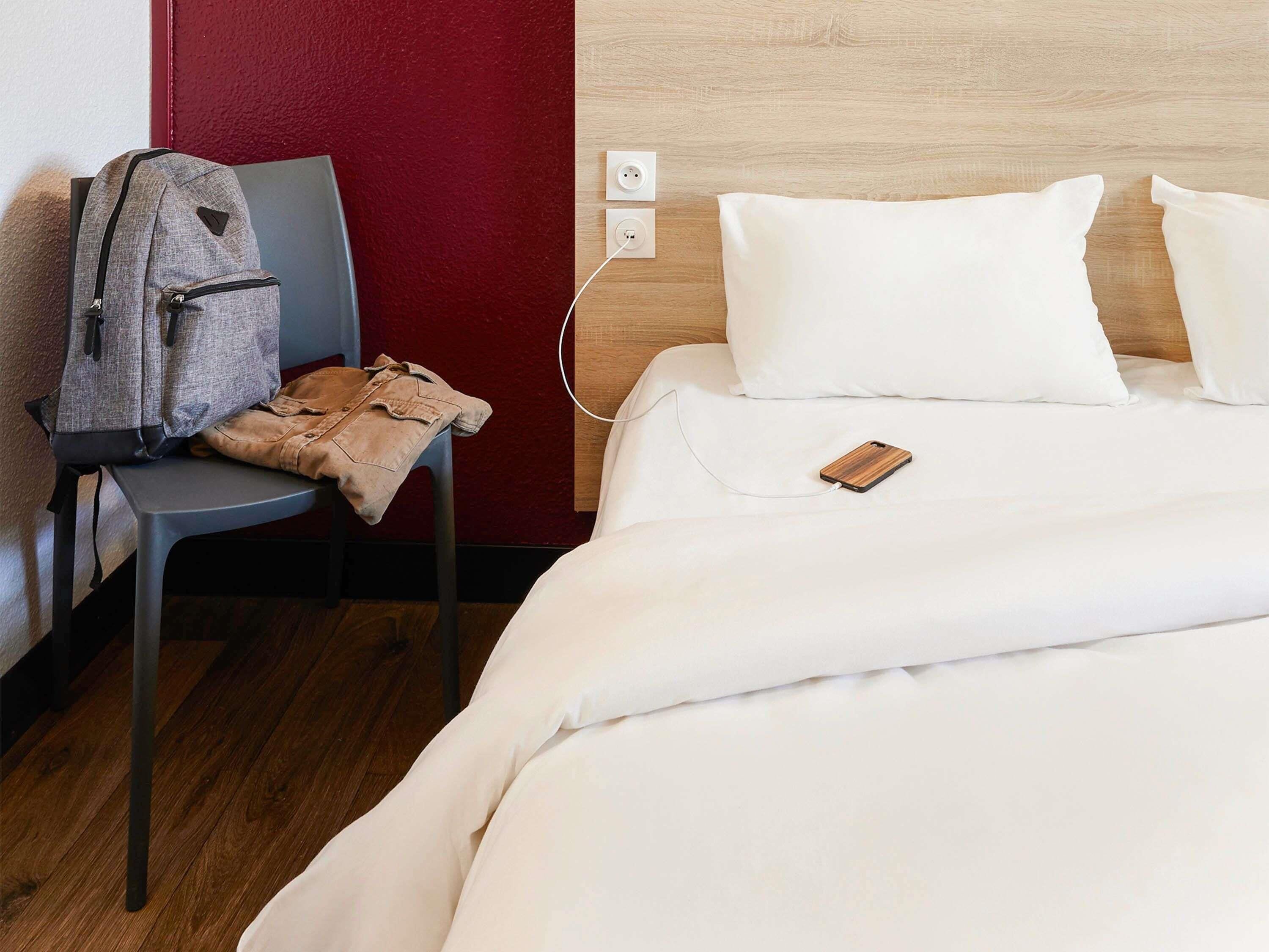 Una habitación de hotel con una cama, una silla, una mochila y una mesita de noche de madera.