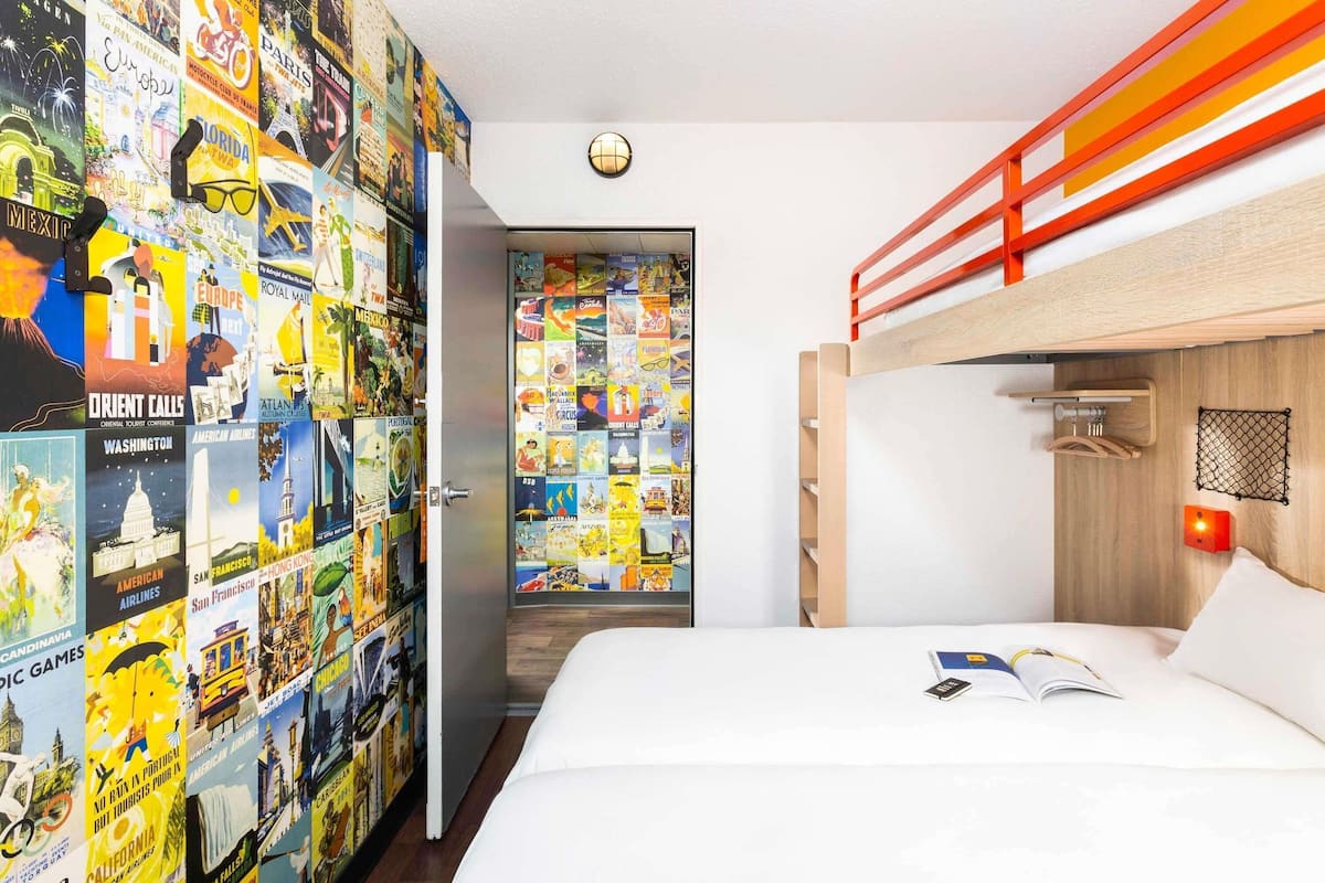 A bedroom with a bunk bed, a wall decorated with travel posters, a door with a round light, and a bedside table with a book and a phone.