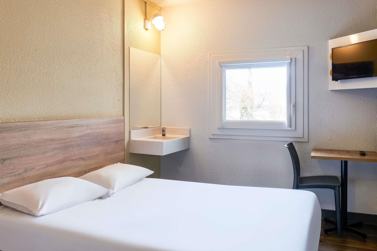 A hotel room with a bed, a bedside table, a mirror, a sink, and a window.