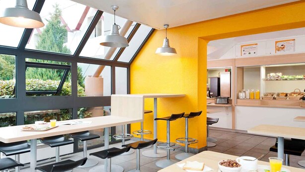 Un café moderne avec des tables hautes et des tabourets de bar, de grandes fenêtres et un mur d’accent jaune vif.