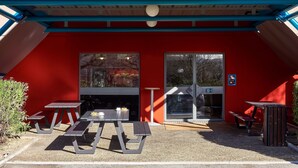 L’entrée d’un hôtel, avec une façade rouge, une enseigne jaune indiquant « hotelFL.com », et un espace extérieur aménagé avec des tables et des bancs.