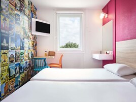 Habitación de hotel con cama, escritorio con silla, televisor montado en la pared y ventana con vistas a follaje.