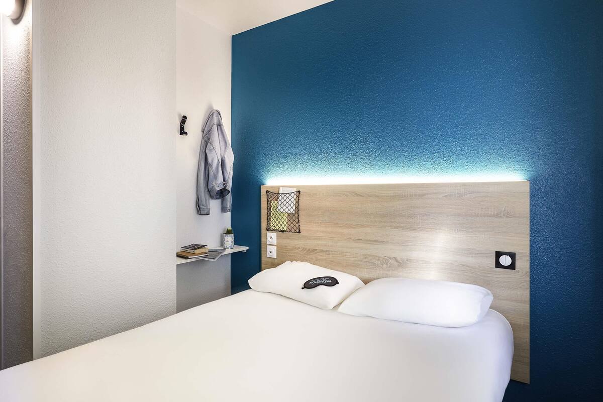 Een hotelkamer met een bed, een hoofdbord met verlichting, een plank met een boek en een plant, en een blauwe accentmuur.