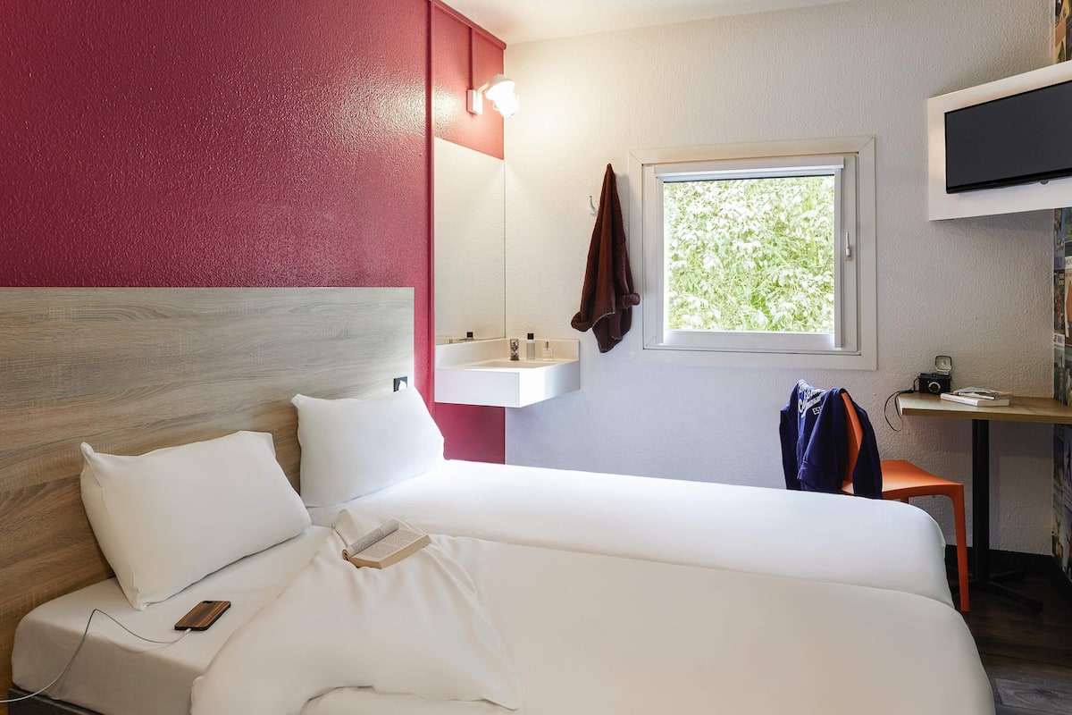 Une chambre d’hôtel avec un lit, une table de chevet avec une lampe, une fenêtre donnant sur un espace verdoyant et un téléviseur fixé au mur.