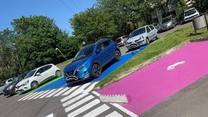 En parkeringsplads med cykelsti i blå og lyserød, afmærkede fodgængerovergange og flere parkerede biler.