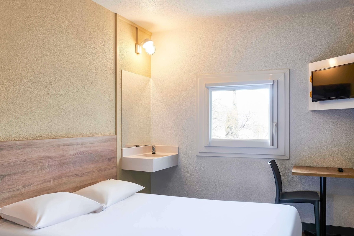 A bedroom with a bed, a bedside table, a mirror, a window, and a wall-mounted TV.