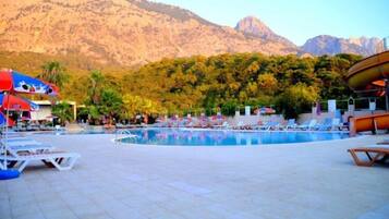 Zona de piscina con sillones de descanso y sombrillas, rodeada de árboles y montañas.