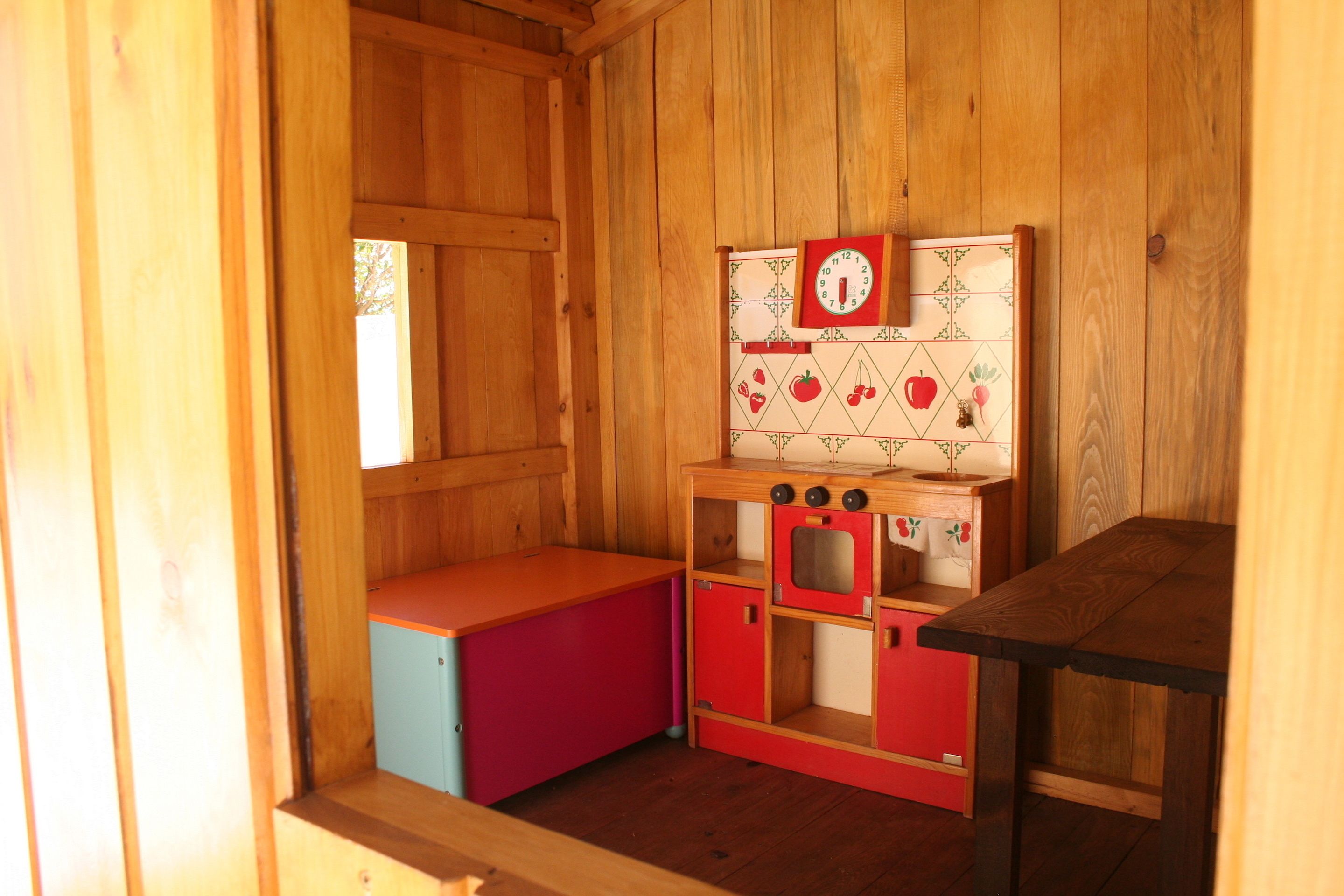 Una habitación de madera con un armario rojo y azul, una pequeña zona de cocina con un reloj y una ventana.