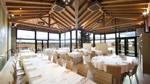 A spacious dining area with high ceilings and large windows offering a view of the landscape. Tables are set with white tablecloths and chairs.