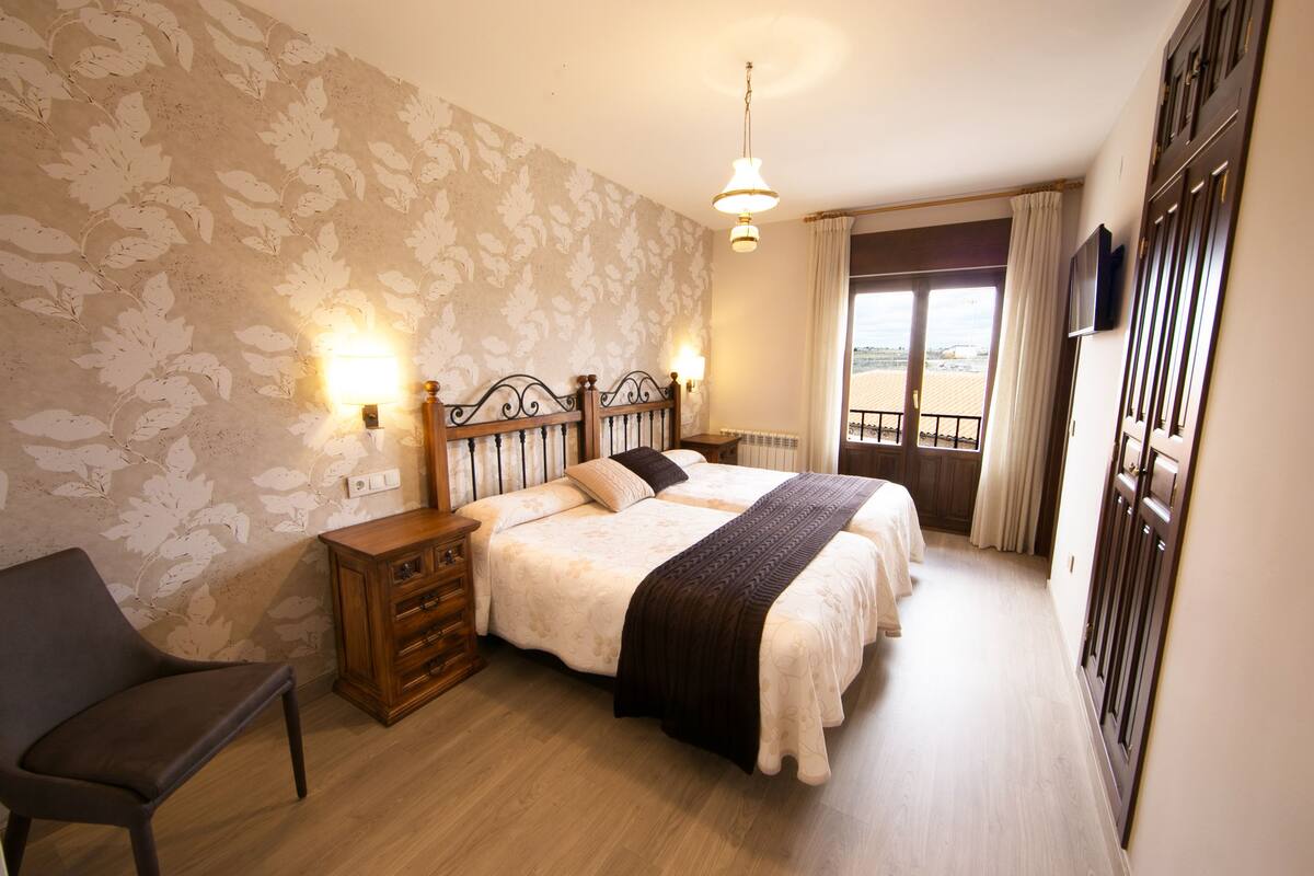 A bedroom with a double bed, a nightstand, a chair, and a view of the beach through the window.