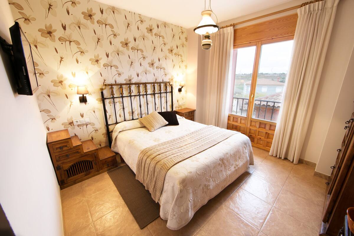 A bedroom with a bed, a nightstand, a lamp, a TV, and a window with curtains.
