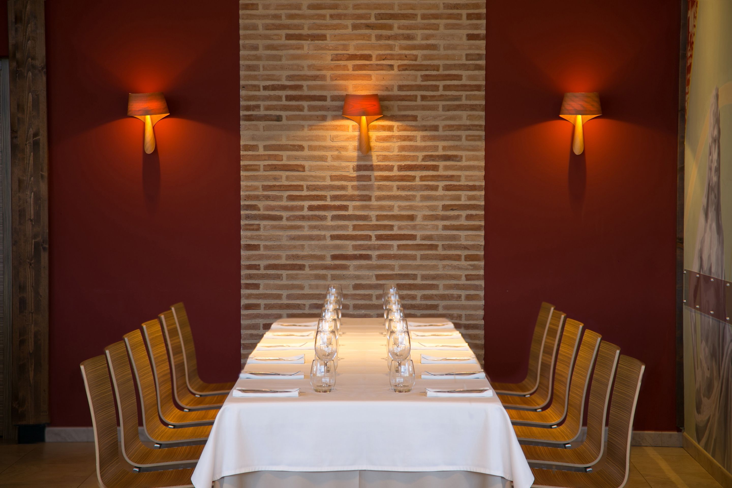 Un comedor con pared de ladrillo, dos lámparas de pared, una mesa larga con sillas y un mantel blanco.