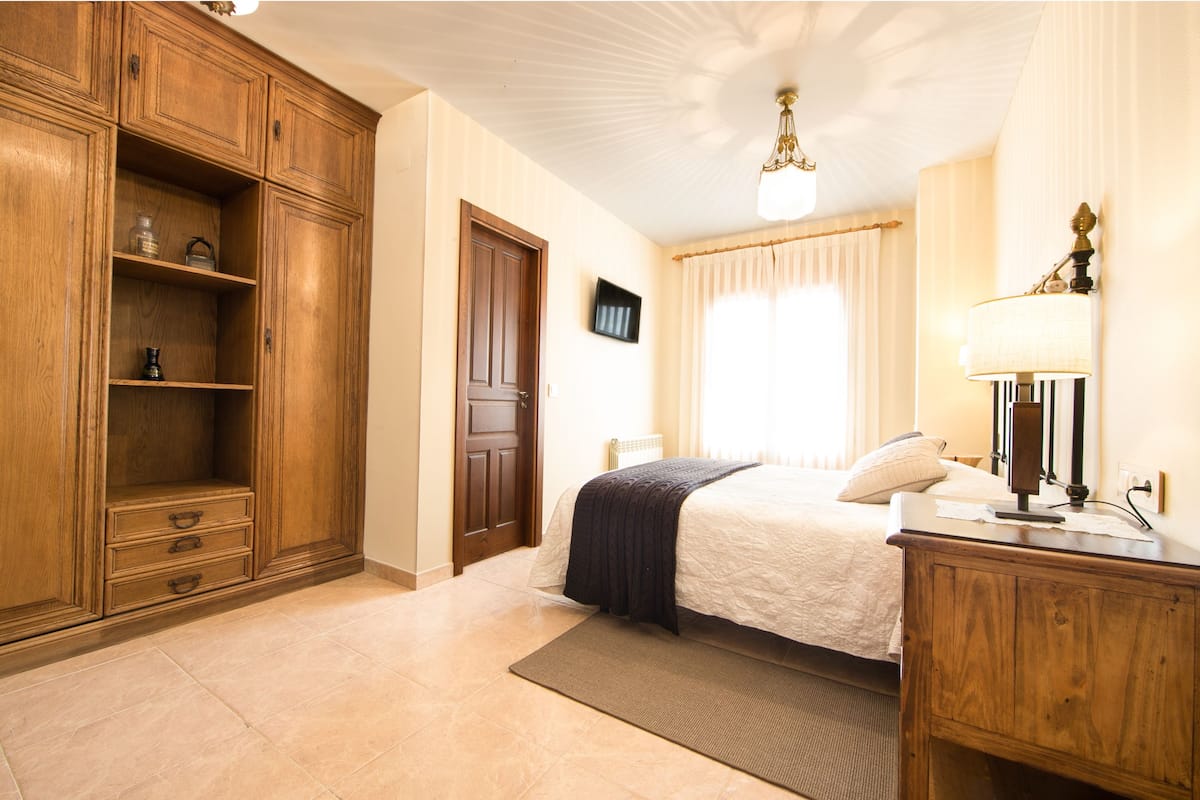 A bedroom with a wooden wardrobe, a bed with a white comforter, a bedside lamp, and a window with curtains.