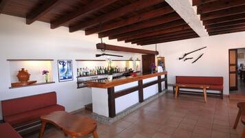Zona de bar con vigas de madera, mostrador con botellas, banco y rifles colgados en la pared.