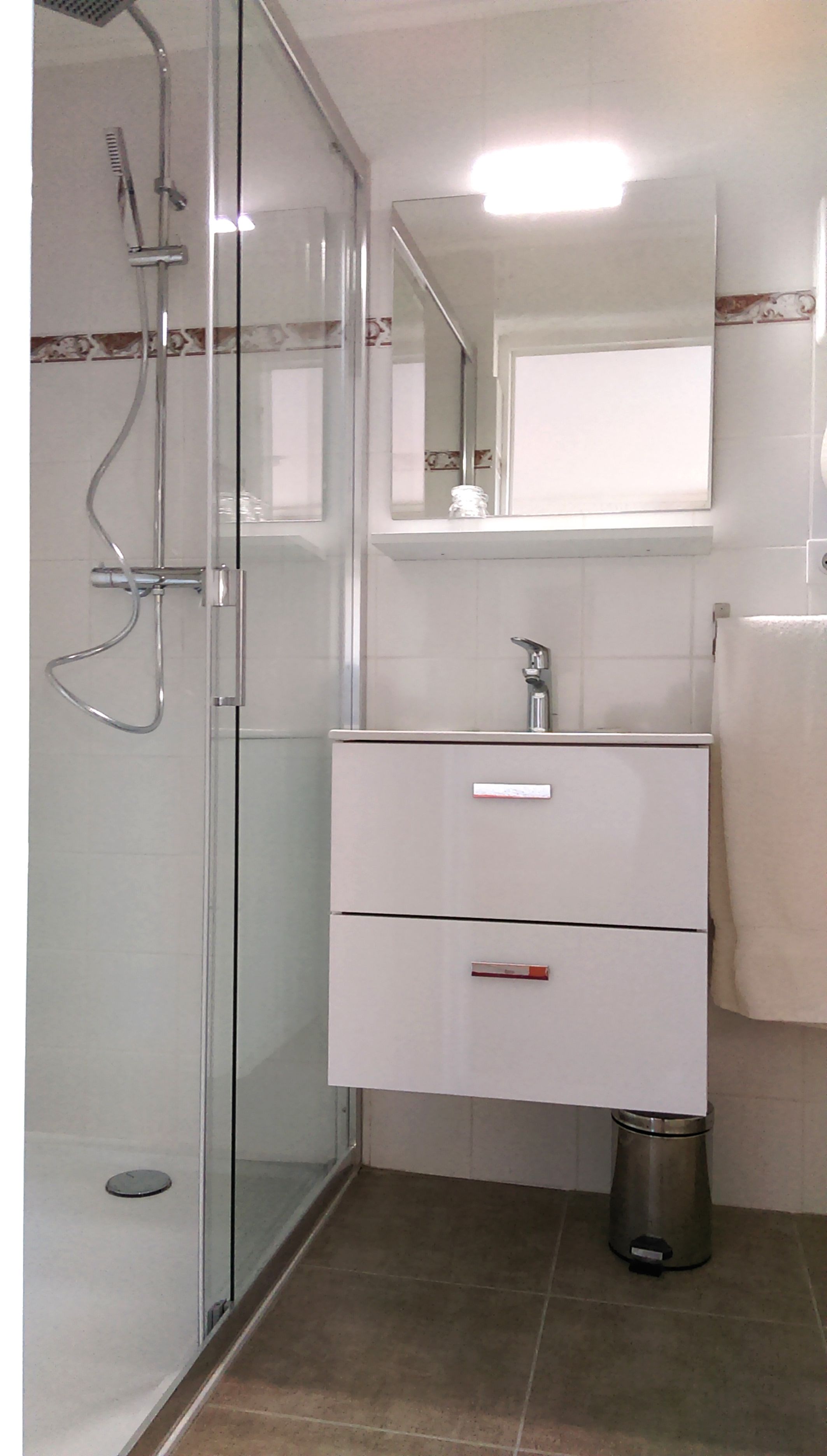 Un baño con cabina de ducha de vidrio, tocador blanco con dos cajones, espejo y toalla colgada en la pared.