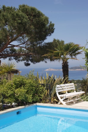 Una piscina con una tumbona blanca, rodeada de follaje y vistas al mar.