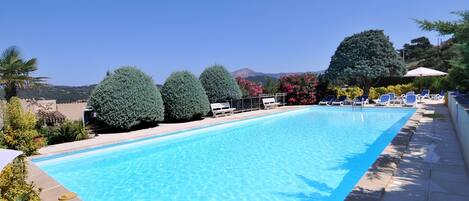A rectangular swimming pool with clear blue water, surrounded by lounge chairs and umbrellas, with a mountain in the background.