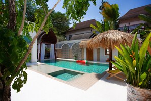 A swimming pool with a thatched roof structure, a white building with a tiled facade, and lush greenery. - Nagawari Art Maldives BBQ Pattaya Villa (Sattahip)