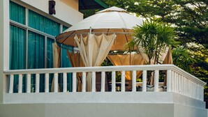 A covered outdoor seating area with a white railing, a beige canopy, and a view of a building with green shutters and a white facade. - Nagawari Art Maldives BBQ Pattaya Villa (Sattahip)