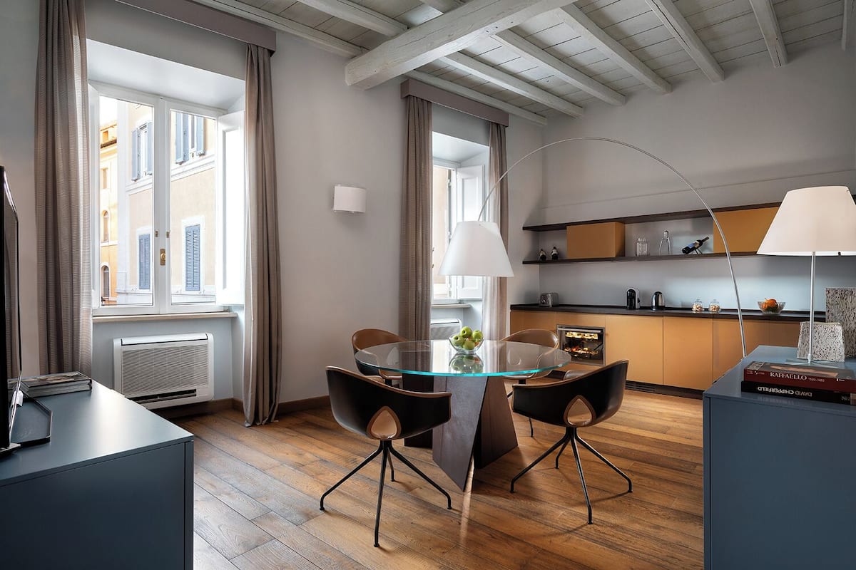 A modern dining area with a glass table, black chairs, and a kitchen in the background.