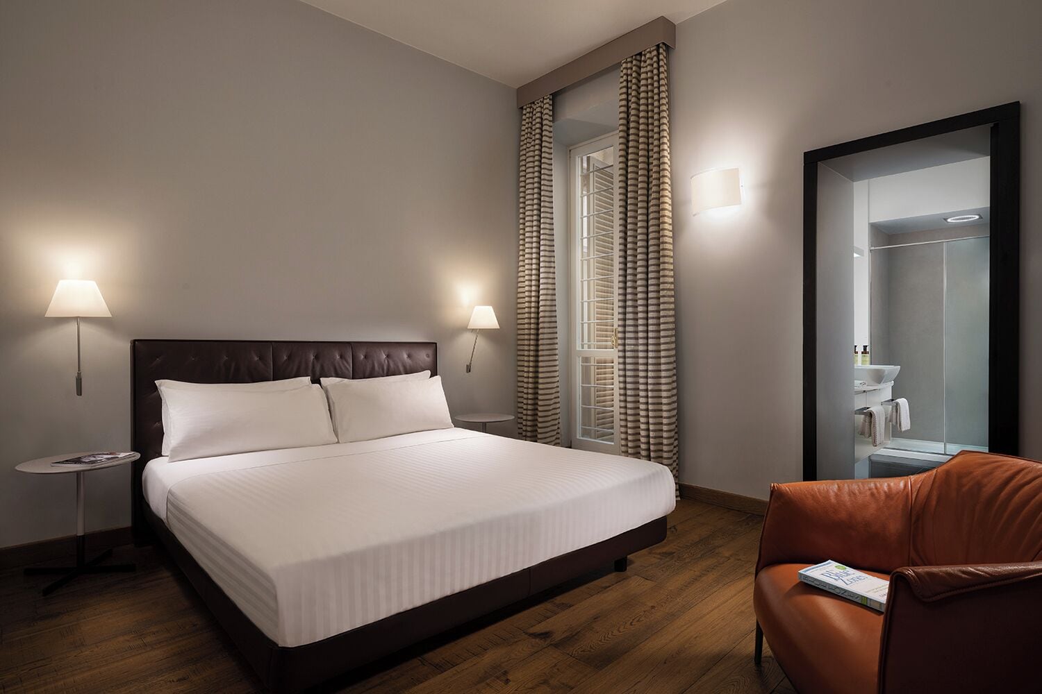 Une chambre d’hôtel avec un grand lit, deux lampes de chevet, une petite table ronde, un fauteuil en cuir et une salle de bain avec un miroir et un lavabo.