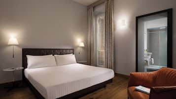 A hotel room with a large bed, two bedside lamps, a small round table, a leather armchair, and a bathroom with a mirror and sink.