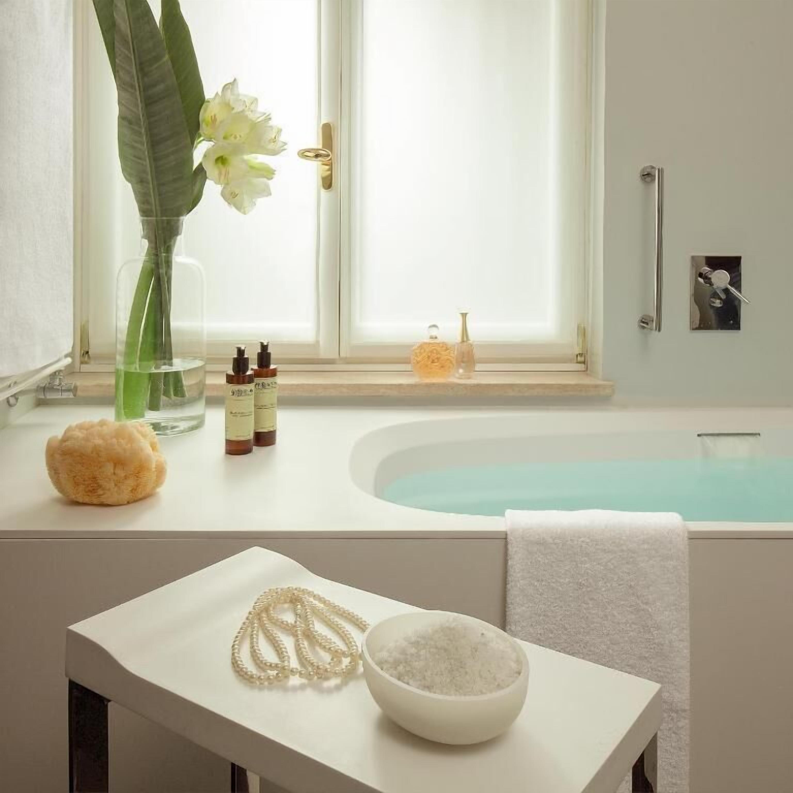 Une salle de bain avec une baignoire, une fenêtre donnant sur un beau paysage, une serviette, un bol de sel et un vase rempli de fleurs.