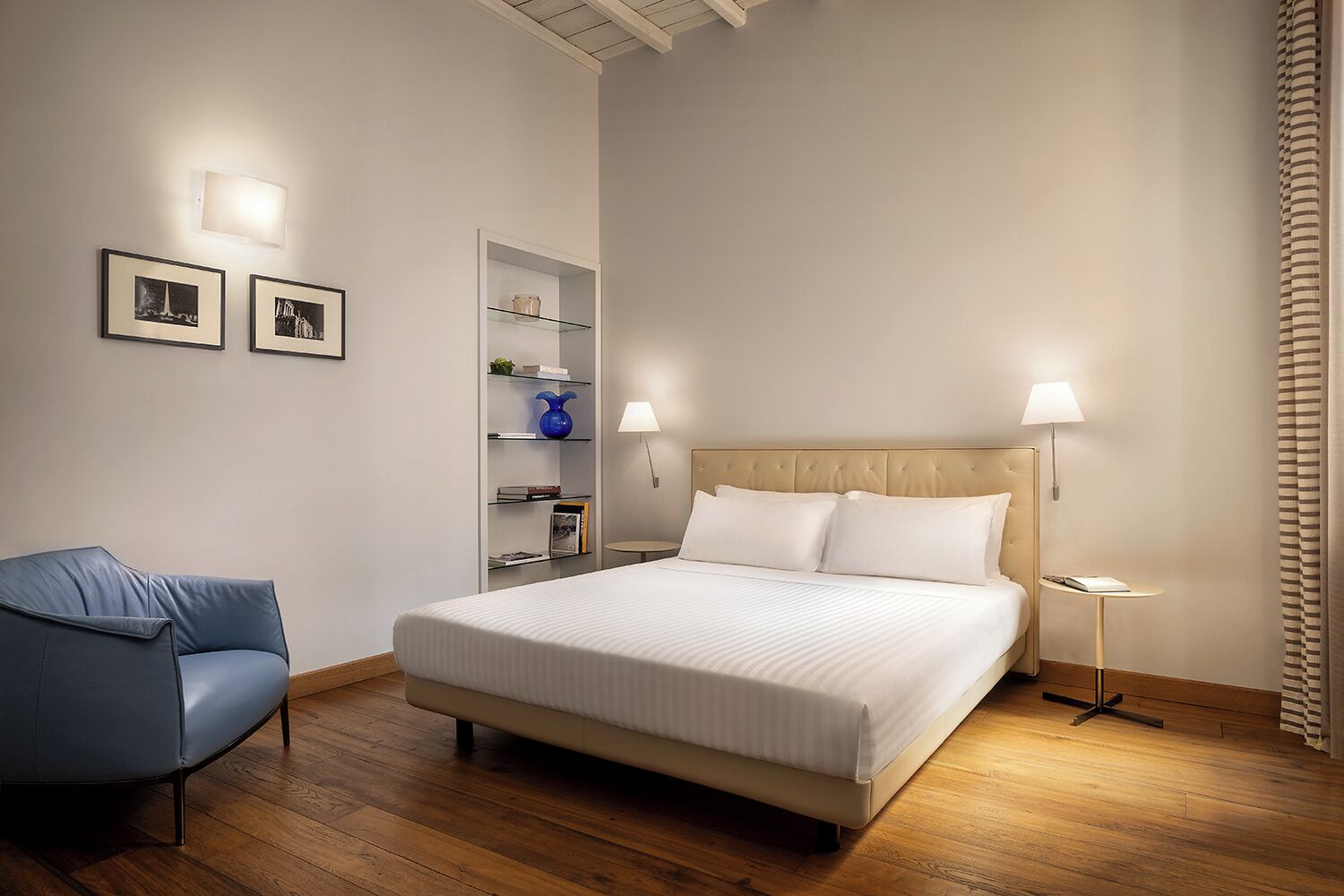 Une chambre d’hôtel avec un grand lit, une chaise bleue, une table de chevet, une lampe et une étagère avec des livres et un vase.