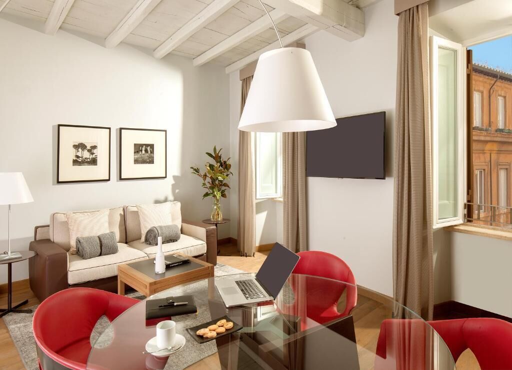 A modern living room with a sofa, red chairs, a glass coffee table with a laptop, a TV, and framed pictures on the wall.