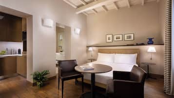 A hotel room with a bed, two chairs, a small table, a nightstand, a lamp, a potted plant, and a kitchen area visible through an open door.
