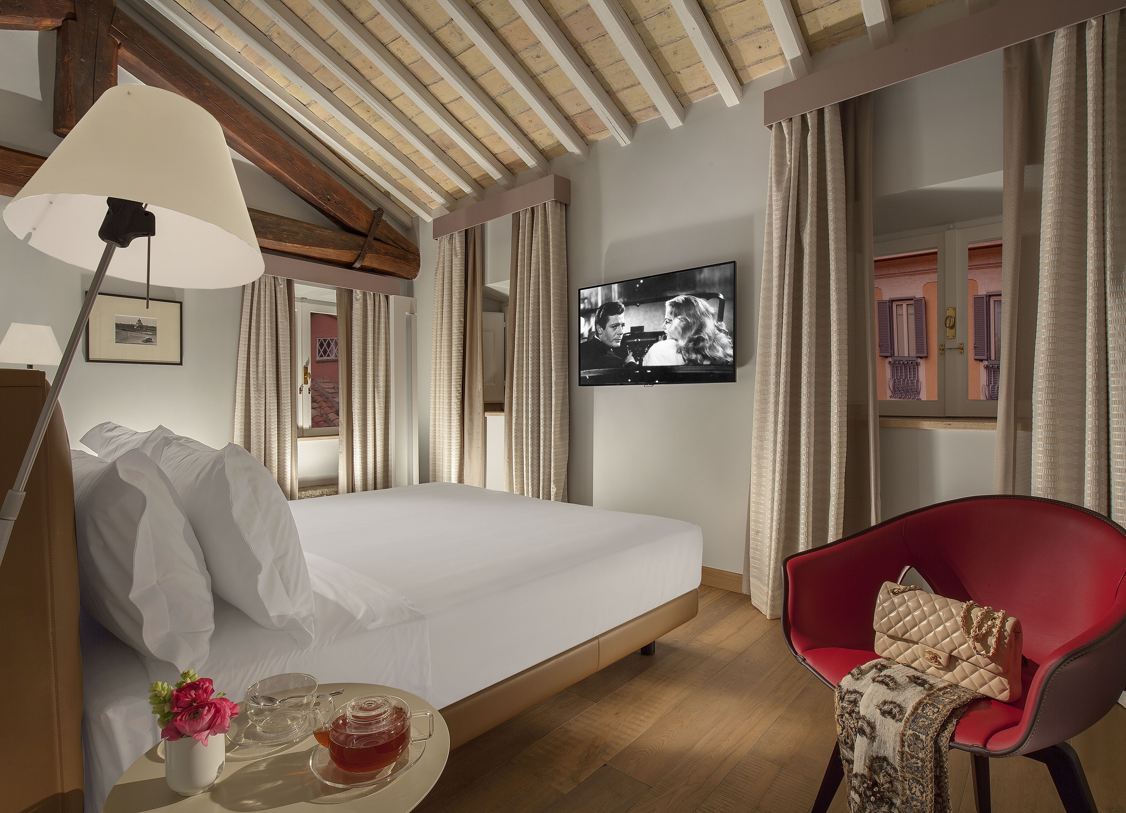 A hotel room with a bed, a red chair, a TV, and a table with a teapot and flowers.