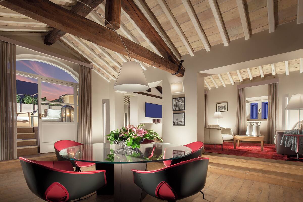 A spacious living room with a glass table and red chairs, wooden beams on the ceiling, and a view of the outdoors.