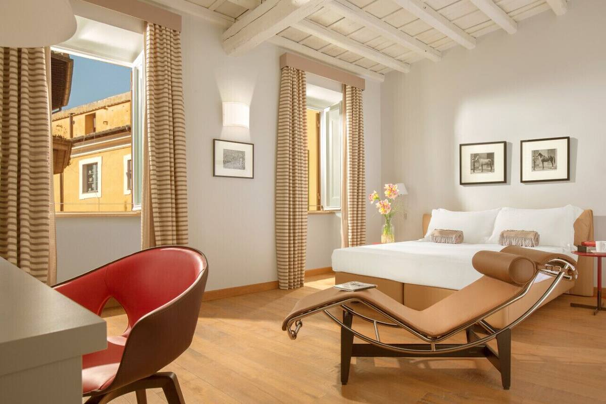 A hotel room with a bed, a red chair, a lounge chair, a small table, and a window with curtains.