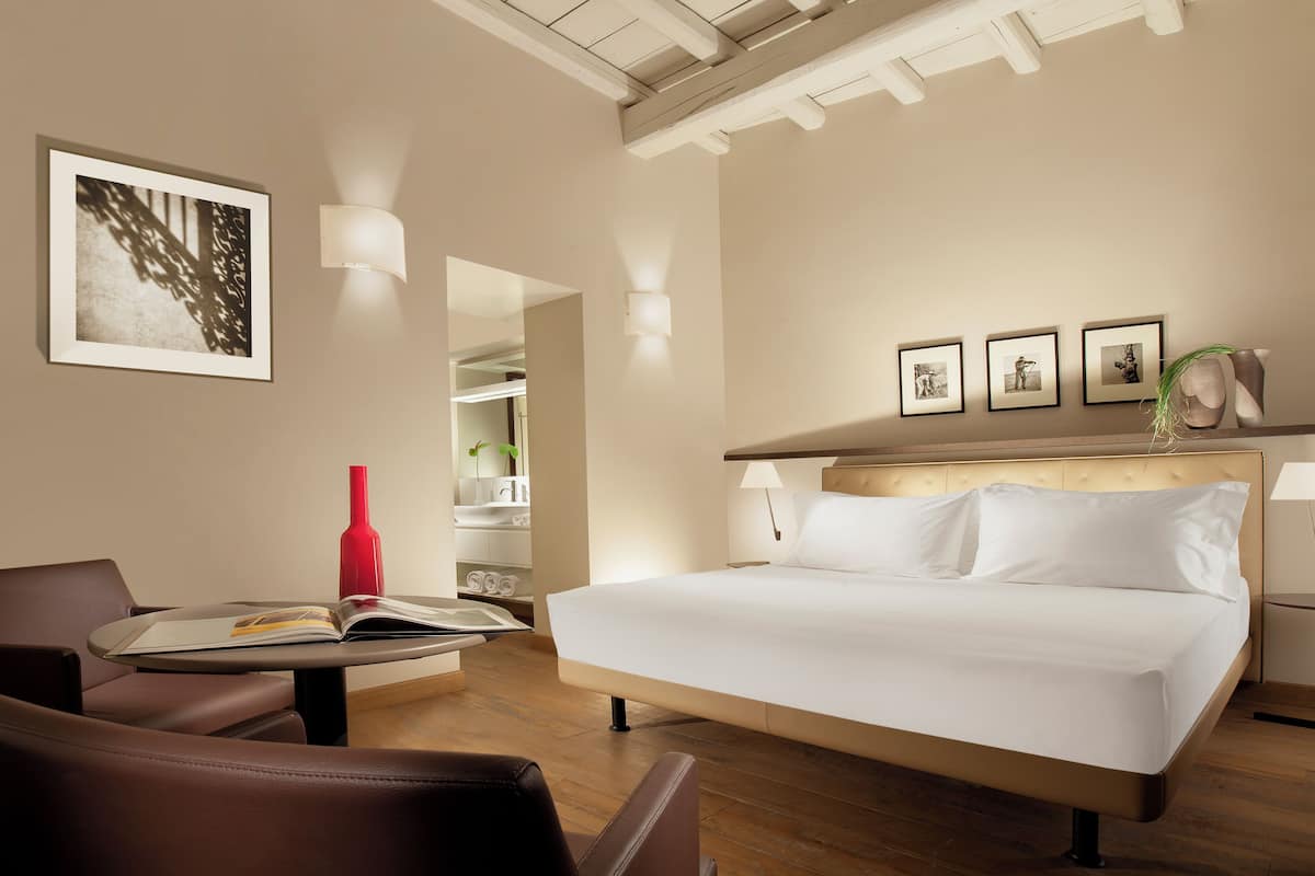 A hotel room with a bed, a chair, a table with a book, and a red vase.