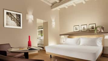 Una habitación de hotel con una cama, una silla, una mesa con un libro y un jarrón rojo.