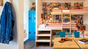 Un lit superposé avec un mur recouvert d’affiches de voyage, un bureau avec un carnet et des lunettes de soleil, et une chaise bleue.