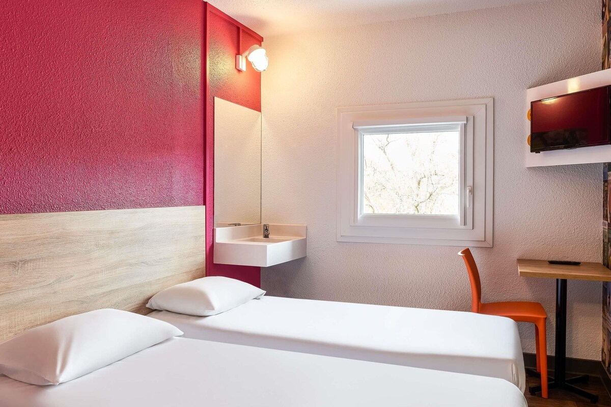 Une chambre d’hôtel moderne avec un lit, un petit bureau, une fenêtre et un mur d’accent rouge.