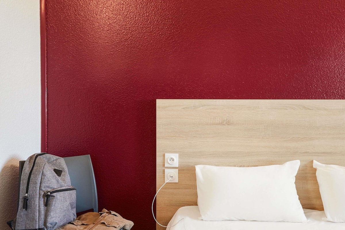 Een hotelkamer met een rode muur, een houten hoofdeinde, een grijze rugzak, een denim jas en een smartphone op het bed.