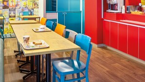 Un espace repas avec une longue table en bois, des chaises bleues et un comptoir rouge où se trouvent une machine à café et divers produits alimentaires.