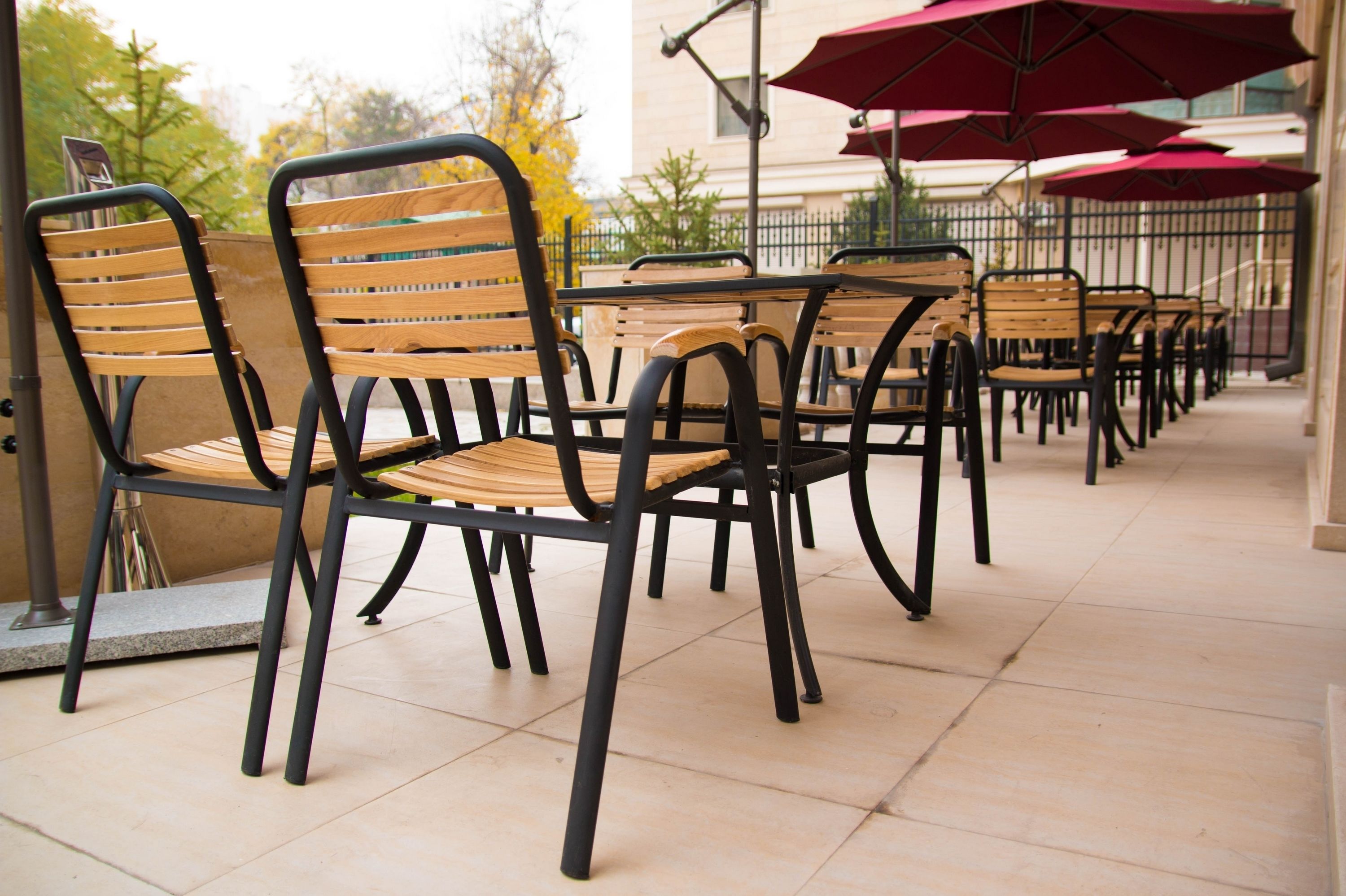 An outdoor seating area with wooden chairs and tables, red umbrellas, and a tiled floor.
