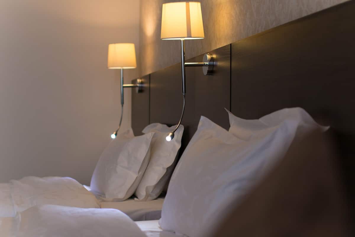 A hotel room with a neatly made bed, two wall-mounted lamps, and a dark headboard.