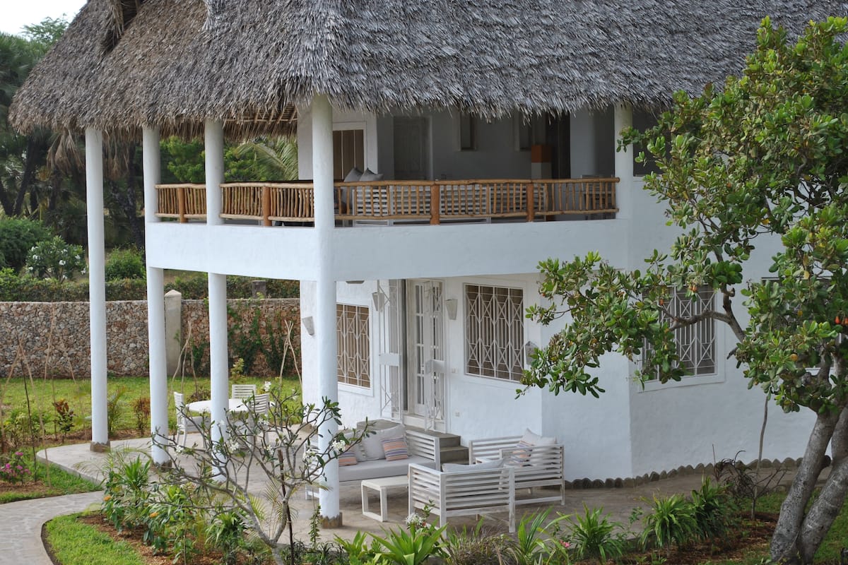 A two-story white building with a thatched roof, a balcony, and a garden area with plants and seating.