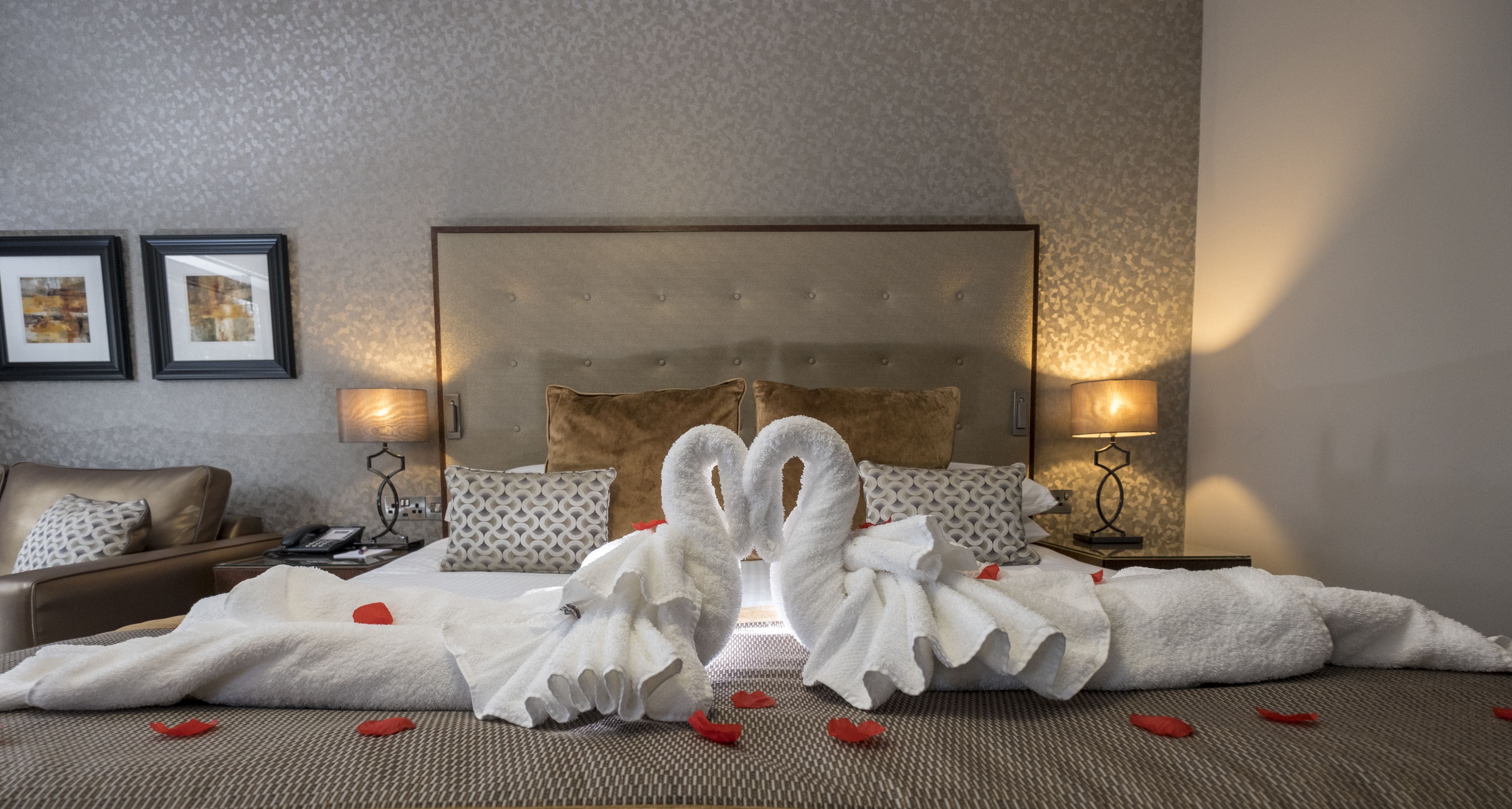 Habitación de hotel con una cama grande, dos lámparas de noche, un sillón de cuero marrón y dos cuadros en la pared.