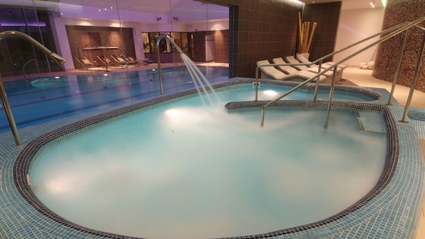 Une piscine intérieure moderne, agrémentée d’une bordure en mosaïque, de chaises longues et d’un élément décoratif aquatique.