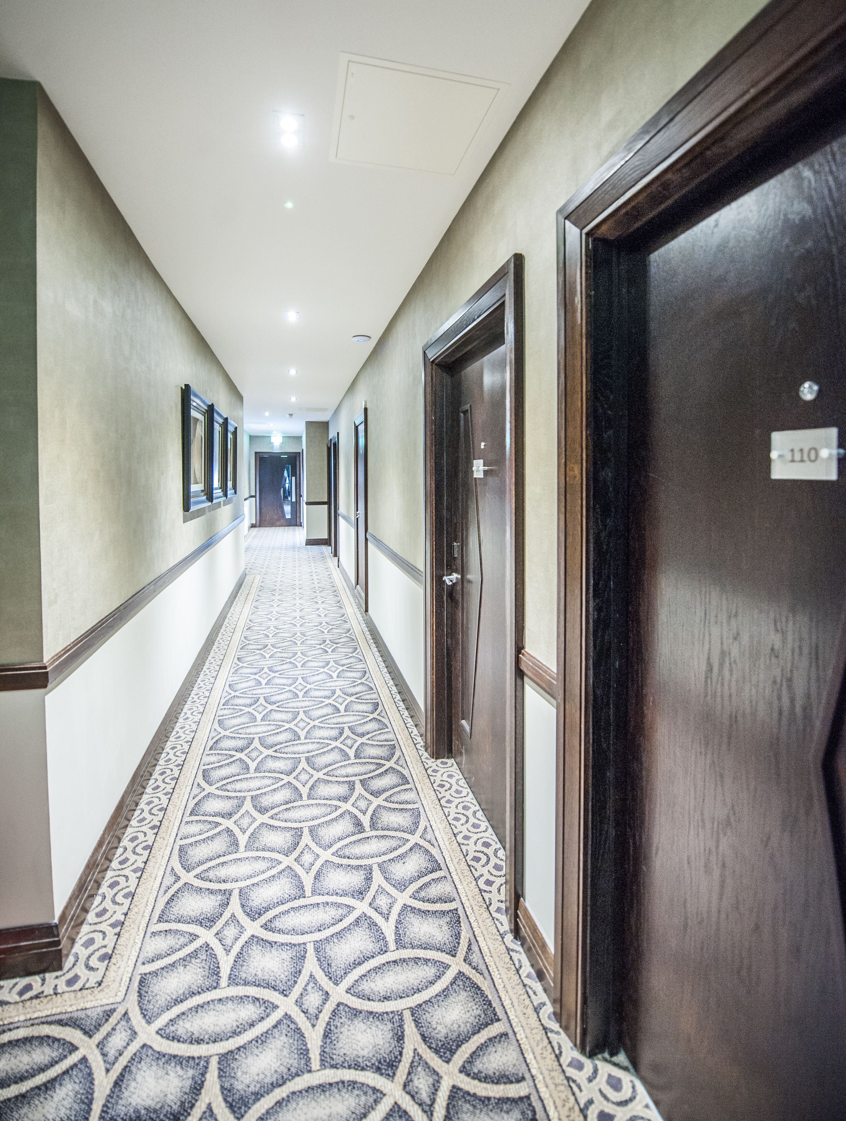 Un pasillo de hotel con alfombra estampada, puertas de madera y cuadros en las paredes.