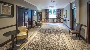 A hotel hallway with patterned carpet, framed artwork on the walls, and a seating area with chairs and a table.
