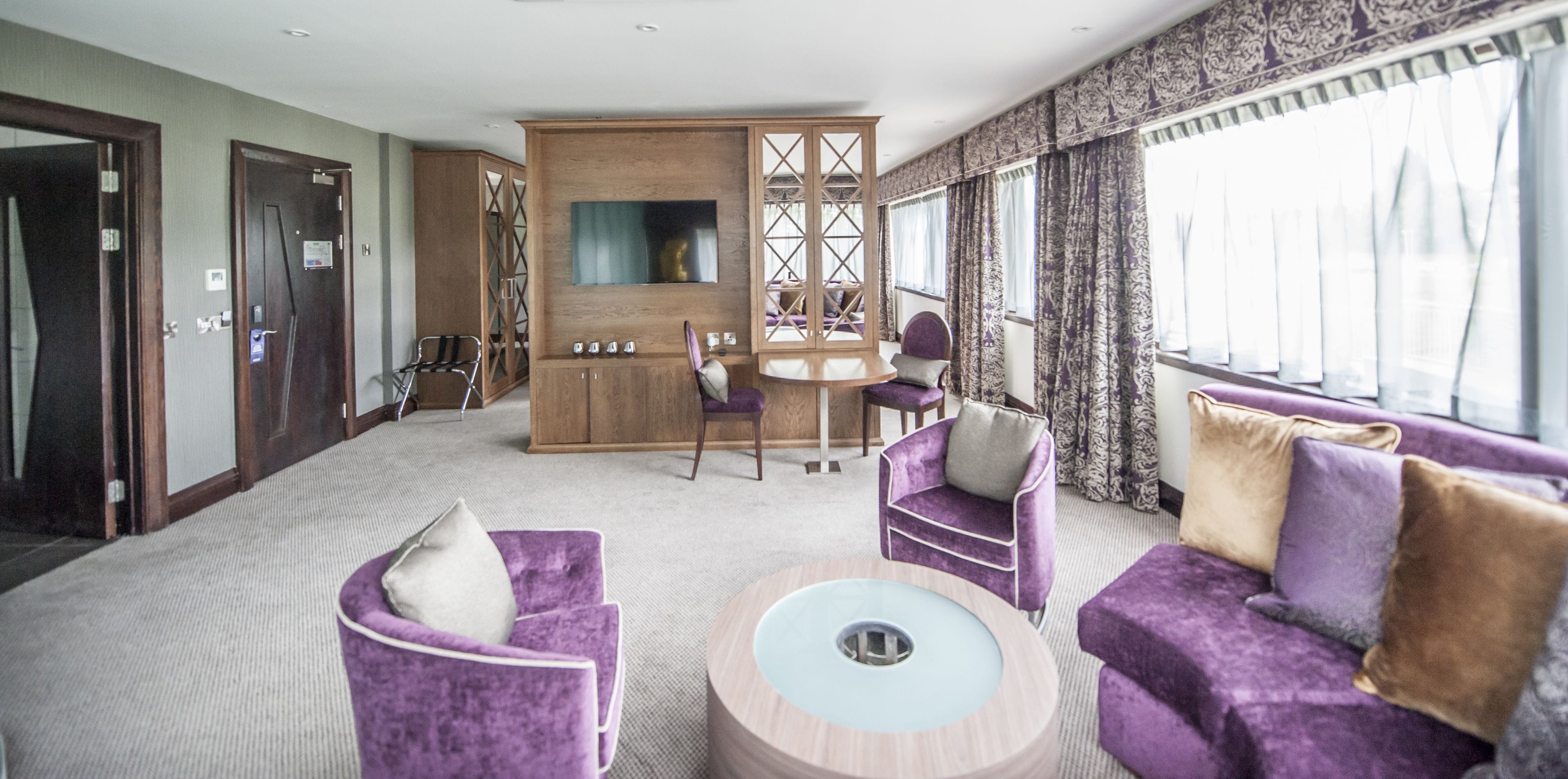A modern hotel room with a seating area, a wooden cabinet, a television, and large windows with curtains.
